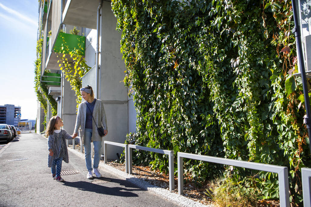 Vanhempi ja lapsi kävelevät viherseinäisen rakennuksen viertä aurinkoisessa säässä